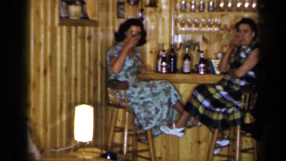 Two women in 1960s dresses sit on stools at a private Long Island home bar, with bottles, glasses, and wood paneling visible around them.