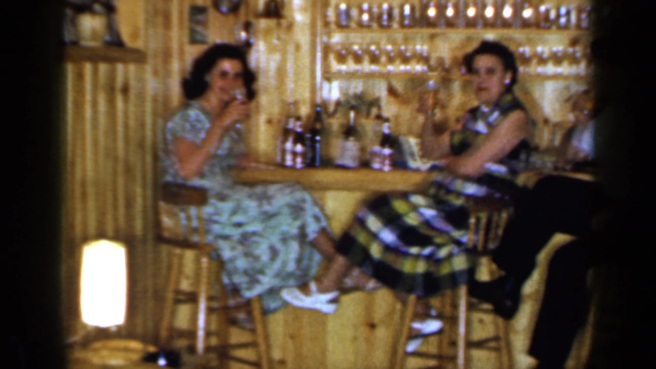Two women sit at a wooden bar with drinks in front of shelves of glasses.