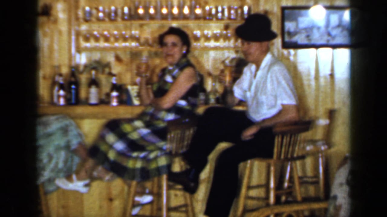 A woman and a man in a dark hat sit at a wooden bar with drinks.