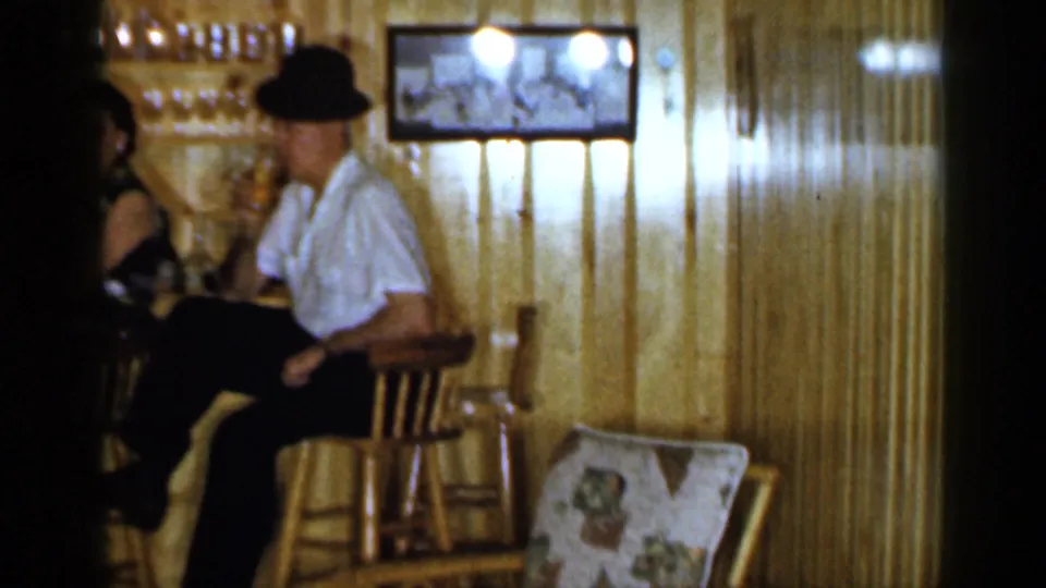 The man in a white shirt and dark hat remains seated at the wooden bar, with wood paneling, stools, and a framed wall picture visible.