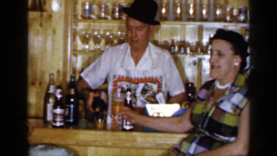 A man in a white shirt and dark hat stands behind a wood-paneled bar as a smiling woman in a plaid dress sits nearby.