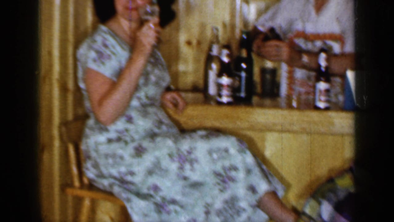 Woman in a floral dress sits at a wood bar holding a drink.
