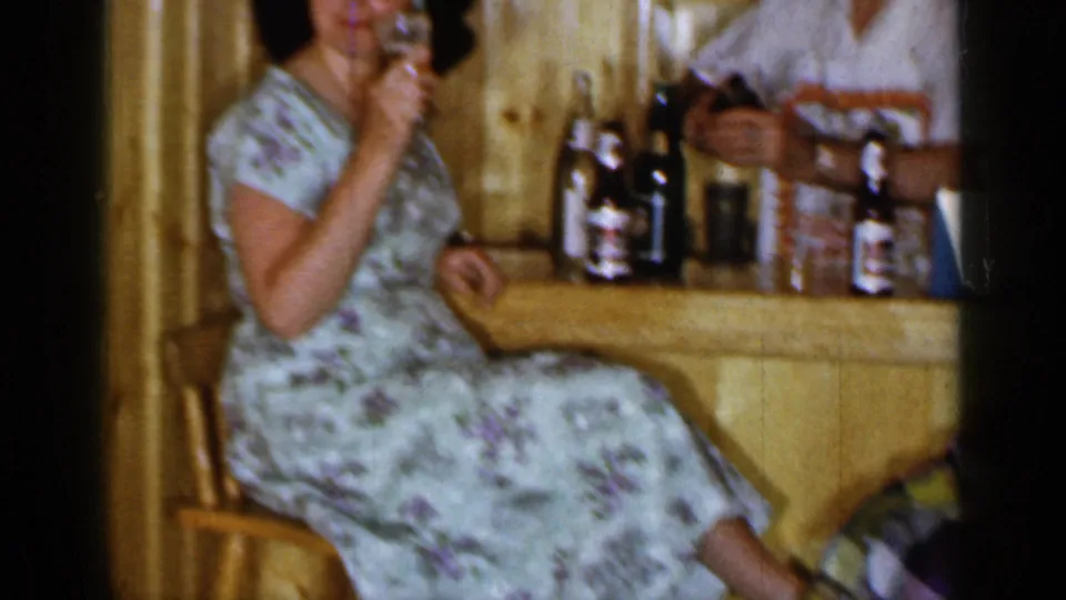 1965 Long Island frame showing a woman in a floral dress seated at a rustic bar, holding a small glass with bottles behind her.