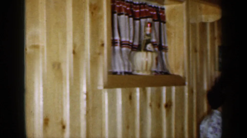 1965 Long Island bar interior detail with pale wood paneling, a small curtained shelf, and part of a seated person at the edge of frame.