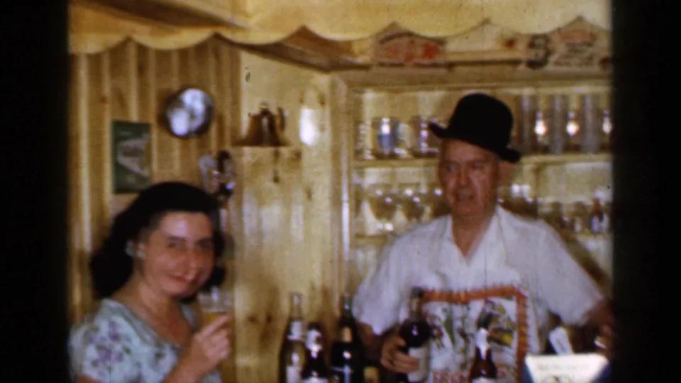 1965 Long Island home-movie frame of a woman holding a drink beside a bartender in a bowler-style hat in a wood-paneled bar.