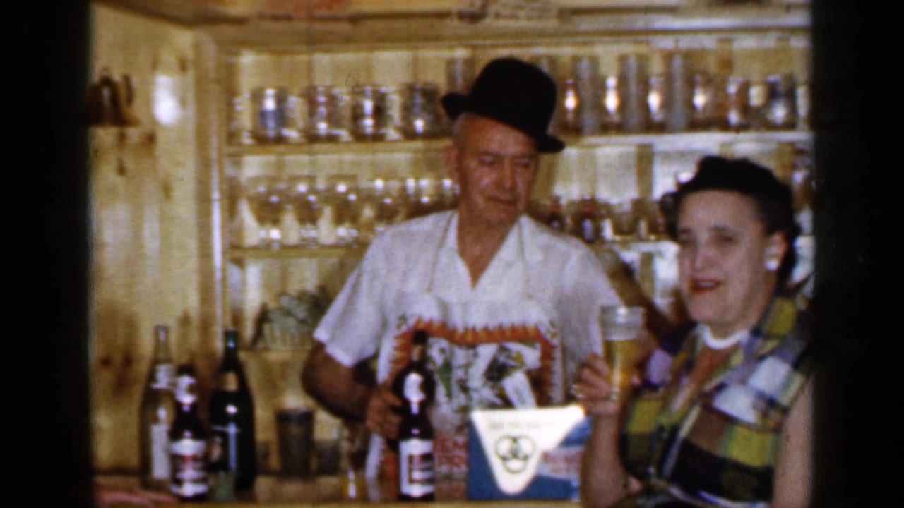 Man in a bowler hat and woman at a wood-paneled bar with beer bottles and glassware.