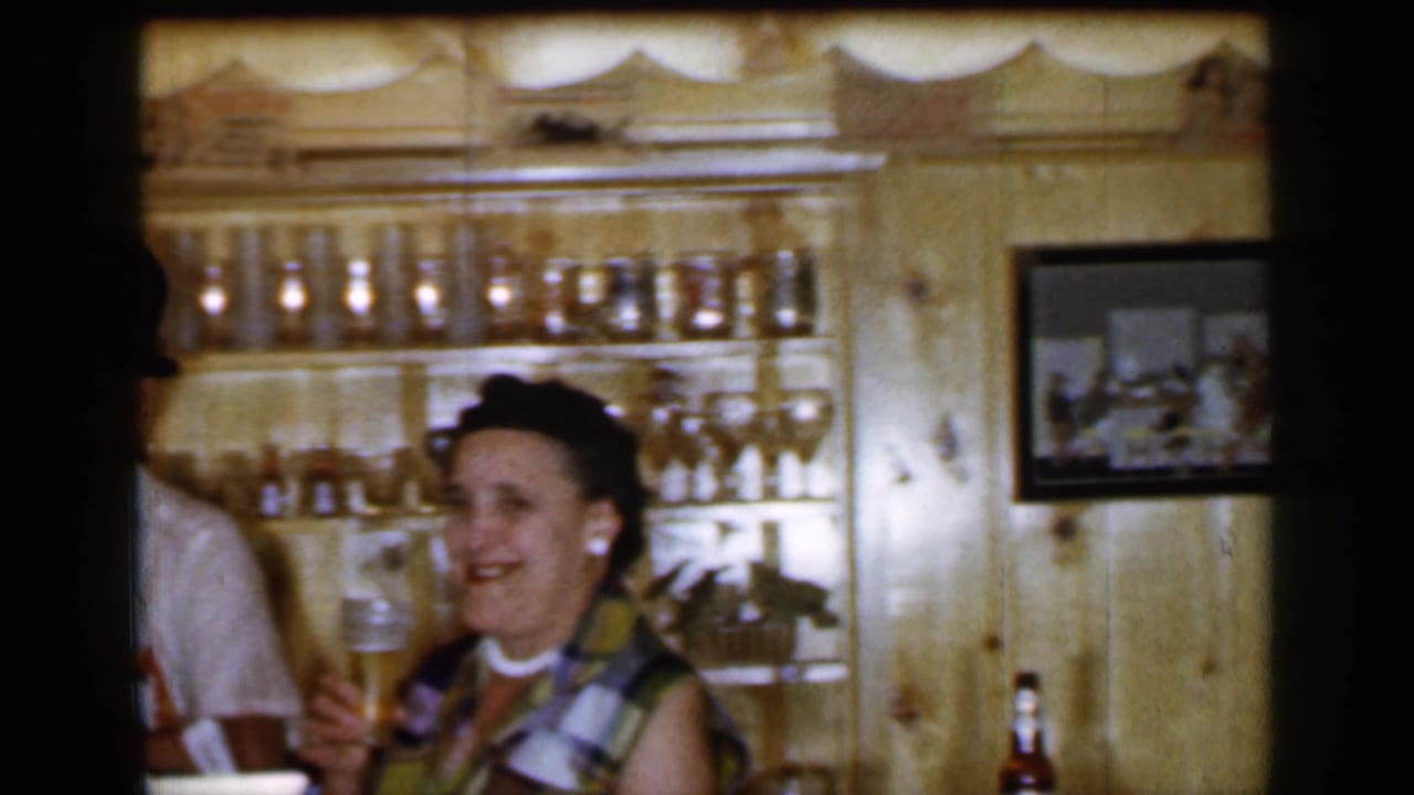 Woman smiling with a beer glass beside shelves of glassware in a wood-paneled bar room.
