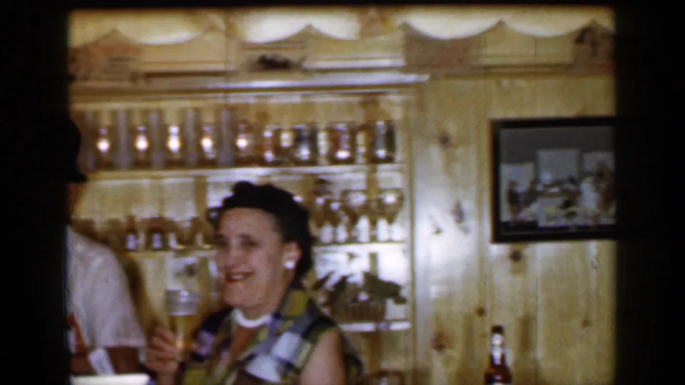 1965 Long Island bar-room frame with an adult woman smiling and holding a beer glass near shelves of glassware and a framed picture.