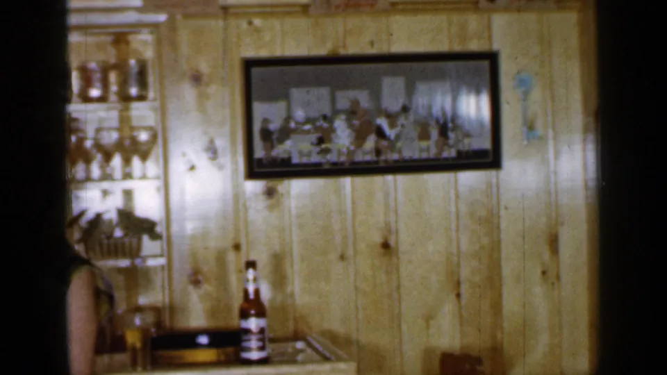 1965 Long Island home-movie frame panning across a bar interior with glassware, framed wall art, bottles, and a partial seated figure.