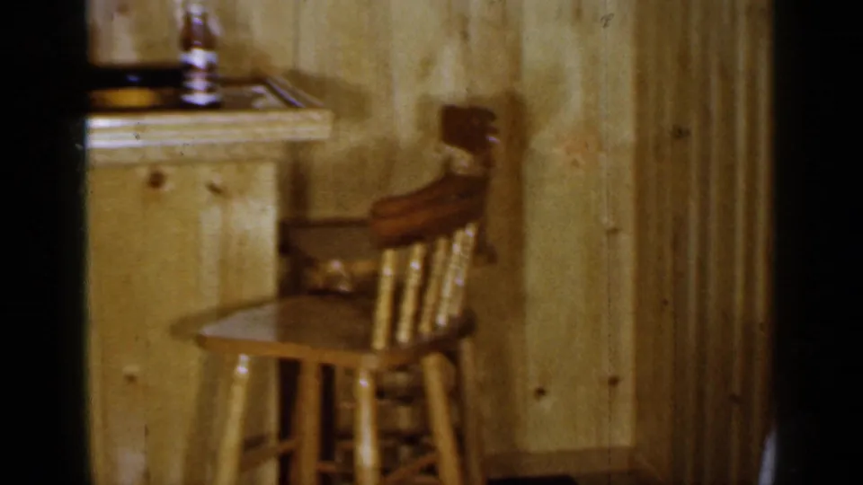 1965 Long Island home-movie frame showing the quieter end of the bar room, with a counter, bottle, wood paneling, and bar stool.
