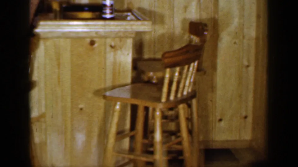 1965 Long Island 8mm frame showing empty wooden stools at a small wood-paneled bar with bottles and glassware nearby.