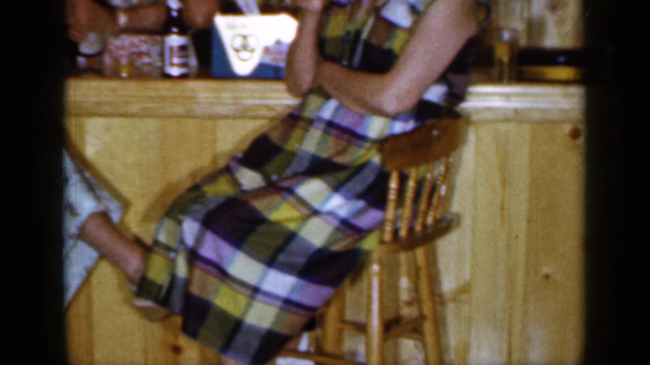 Woman in a plaid dress seated on a wooden stool at a bar counter.