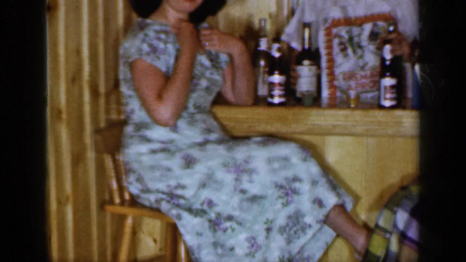 1965 Long Island 8mm frame showing a woman in a floral dress seated at a wood-paneled bar with bottles and glassware.