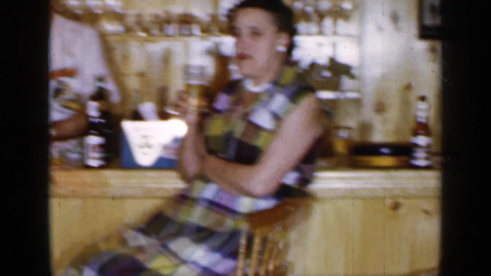 1965 Long Island frame with a seated woman in a plaid dress and a bartender behind a wood-paneled bar with bottles and shelves.
