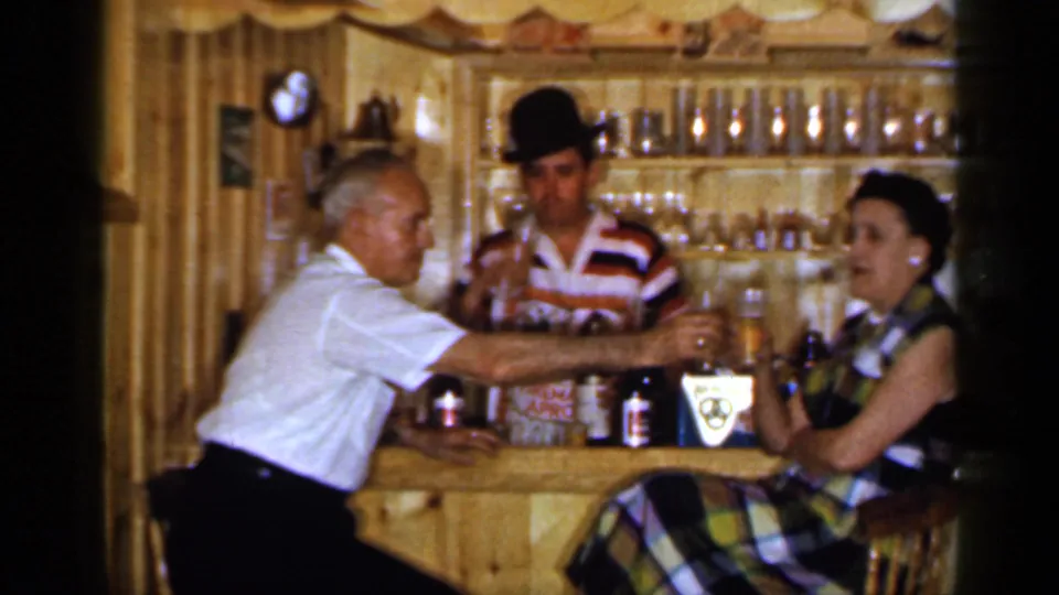 1965 Long Island bar interior with an older man extending a glass toward a seated woman as the bartender stands behind the counter.