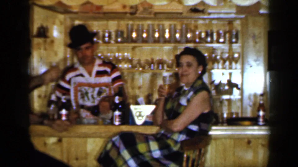 1965 Long Island home-movie frame showing a woman in a plaid dress holding a drink near a bartender in a striped shirt.