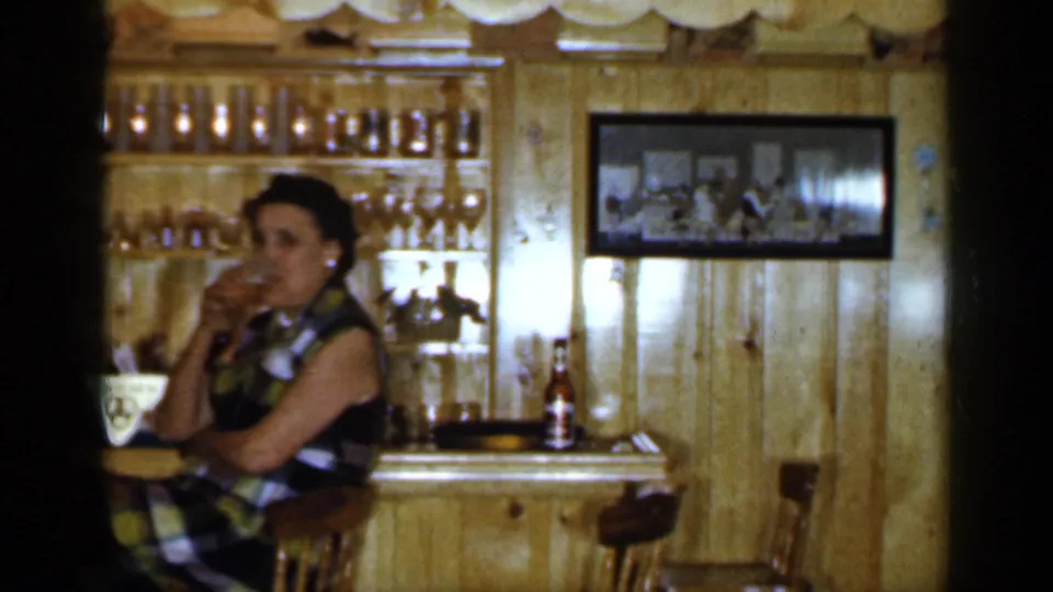 1965 Long Island interior showing a woman taking a drink at a wood-paneled bar with glass shelves and a framed wall picture.