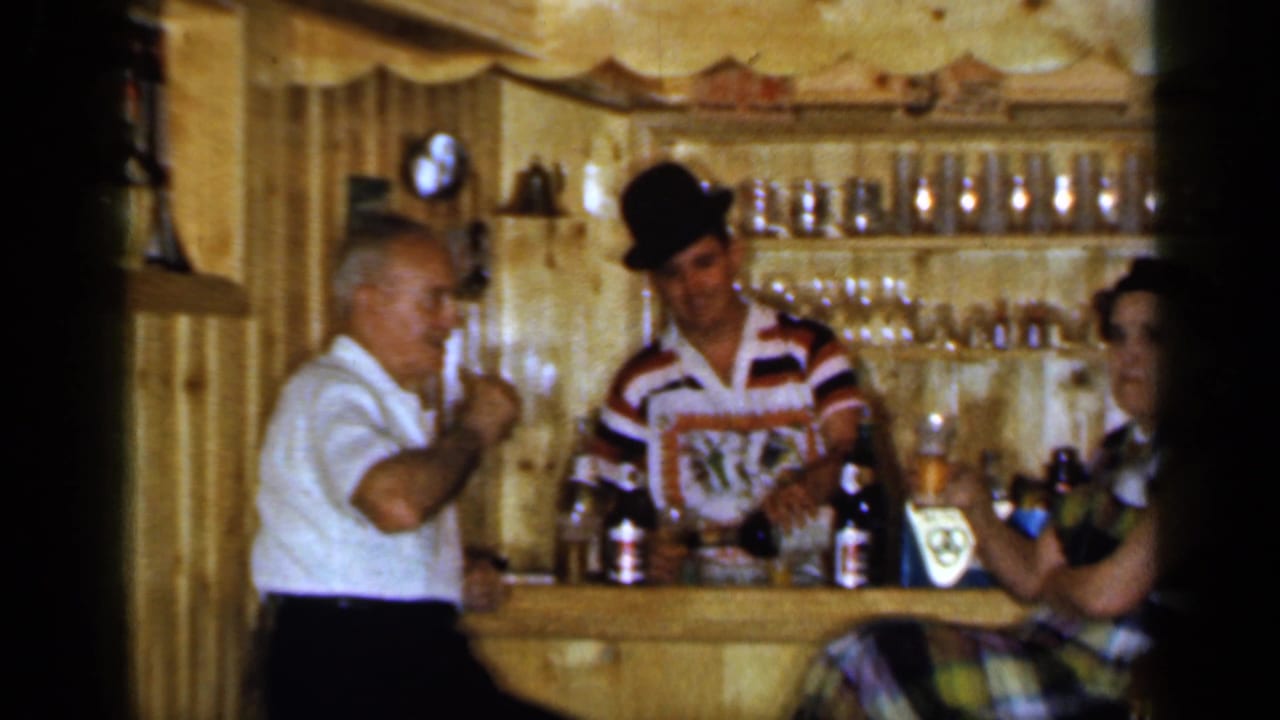 Older man raises a glass beside a bartender and seated woman at a tavern bar.