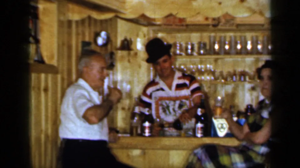 Inside the 1965 Long Island tavern, an older man raises a glass near the bartender while a woman sits at the bar.