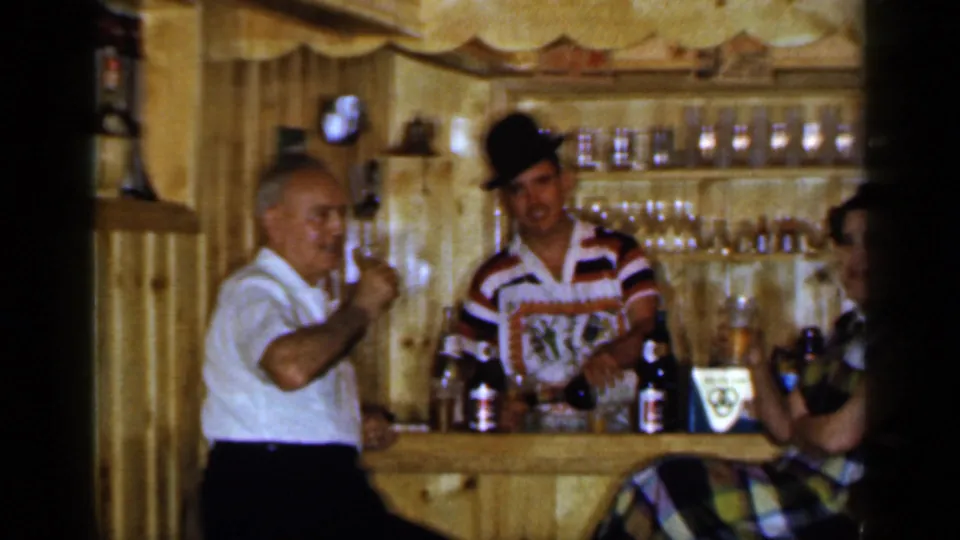 1965 Long Island bar scene with an older man toasting, a bartender in a striped shirt, and a seated woman beside bottles and glassware.