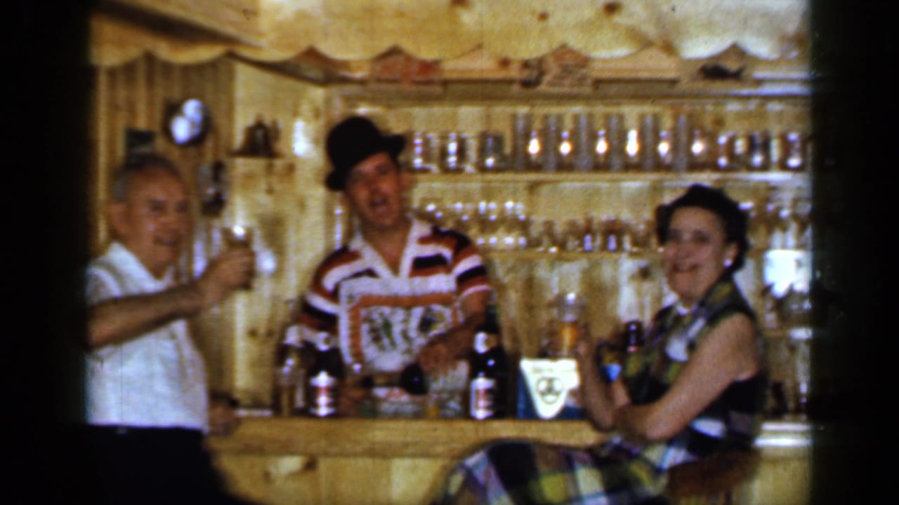 Three adults at a small wooden bar raising drinks inside a wood-paneled room.