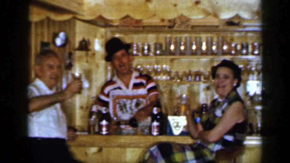 1965 Long Island home-movie frame of three adults smiling and raising drinks at a rustic yellow wooden bar.