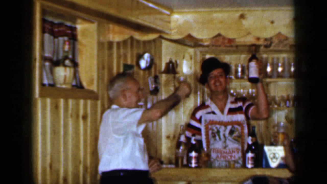 1965 Long Island home-movie frame of two men raising drinks at a small wood-paneled bar with bottles visible.
