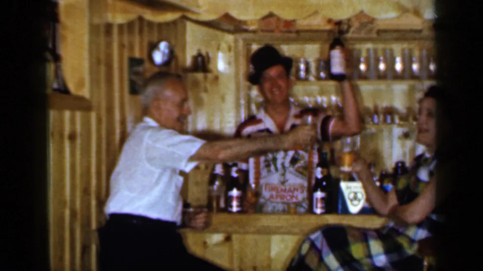 1965 Long Island home-movie frame of three adults mid-toast in a rustic wood-paneled barroom.