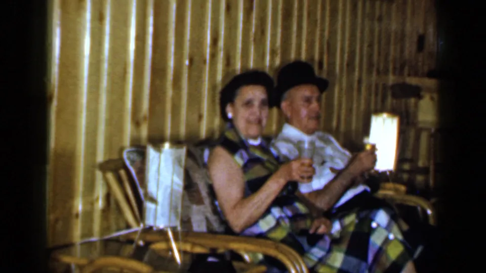 1965 Long Island home-movie frame of a seated woman and man holding drinks beside a lamp in a wood-paneled interior.