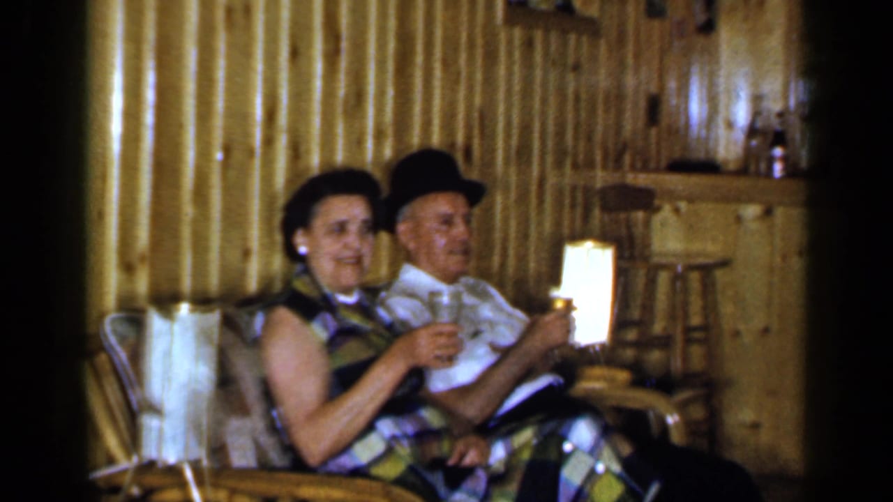 Two adults sit beside a lamp in a wood-paneled bar room holding drinks.