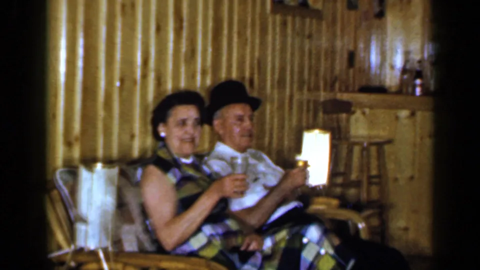 In a 1965 Long Island home movie, a woman and man sit in a wood-paneled club room holding drinks near a glowing lamp.