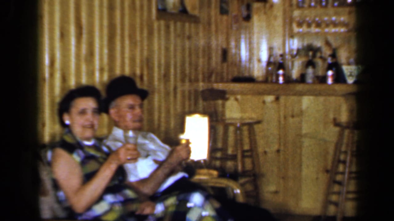 A seated couple holds drinks with a bar counter and stools behind them.