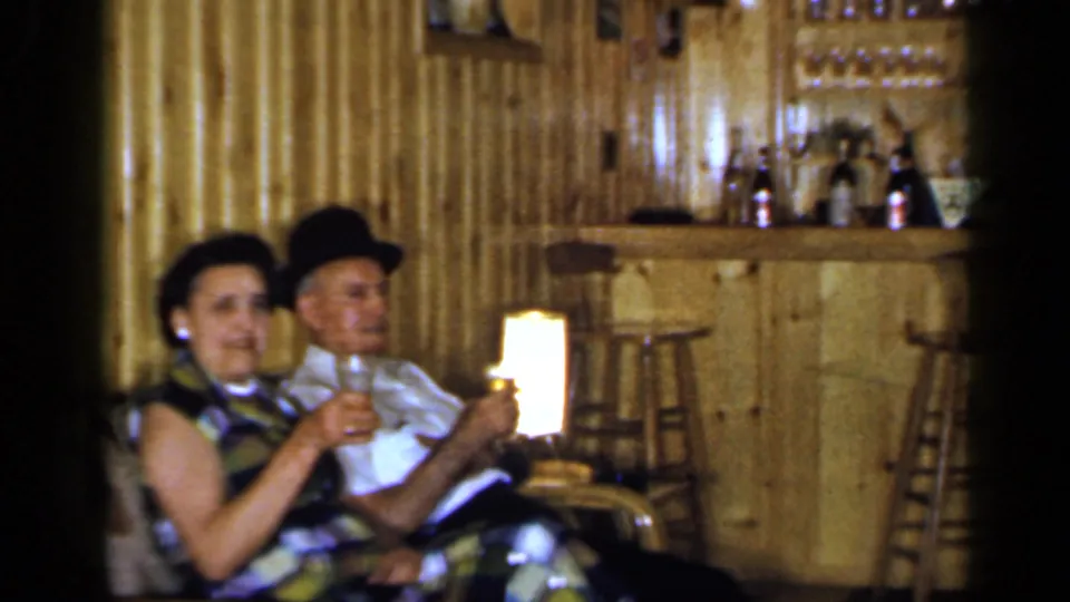 The couple remains seated with drinks in a wood-paneled Long Island club room, with bar stools and shelves in the background.