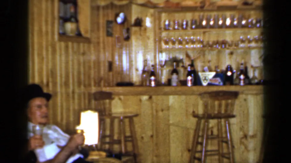 The handheld view pans across the 1965 wood-paneled club room, showing the bar, bottles, hanging glasses, stools, and part of the seated man.