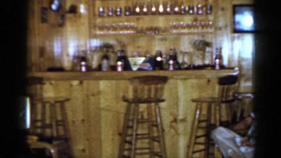 The 1965 Long Island club room view shows a wooden bar, bottles, stools, shelves of glasses, a small television, and part of a seated person.
