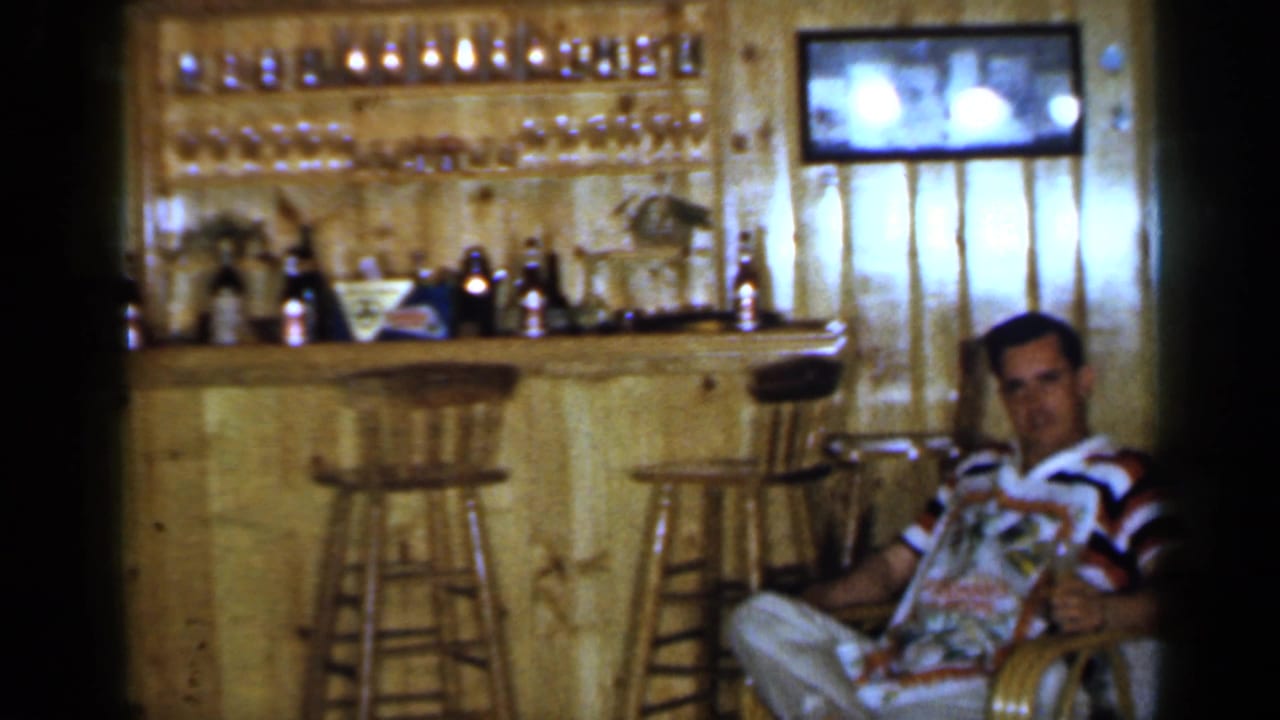 Man in a striped shirt sits near a wood-paneled bar with stools and bottles behind him.