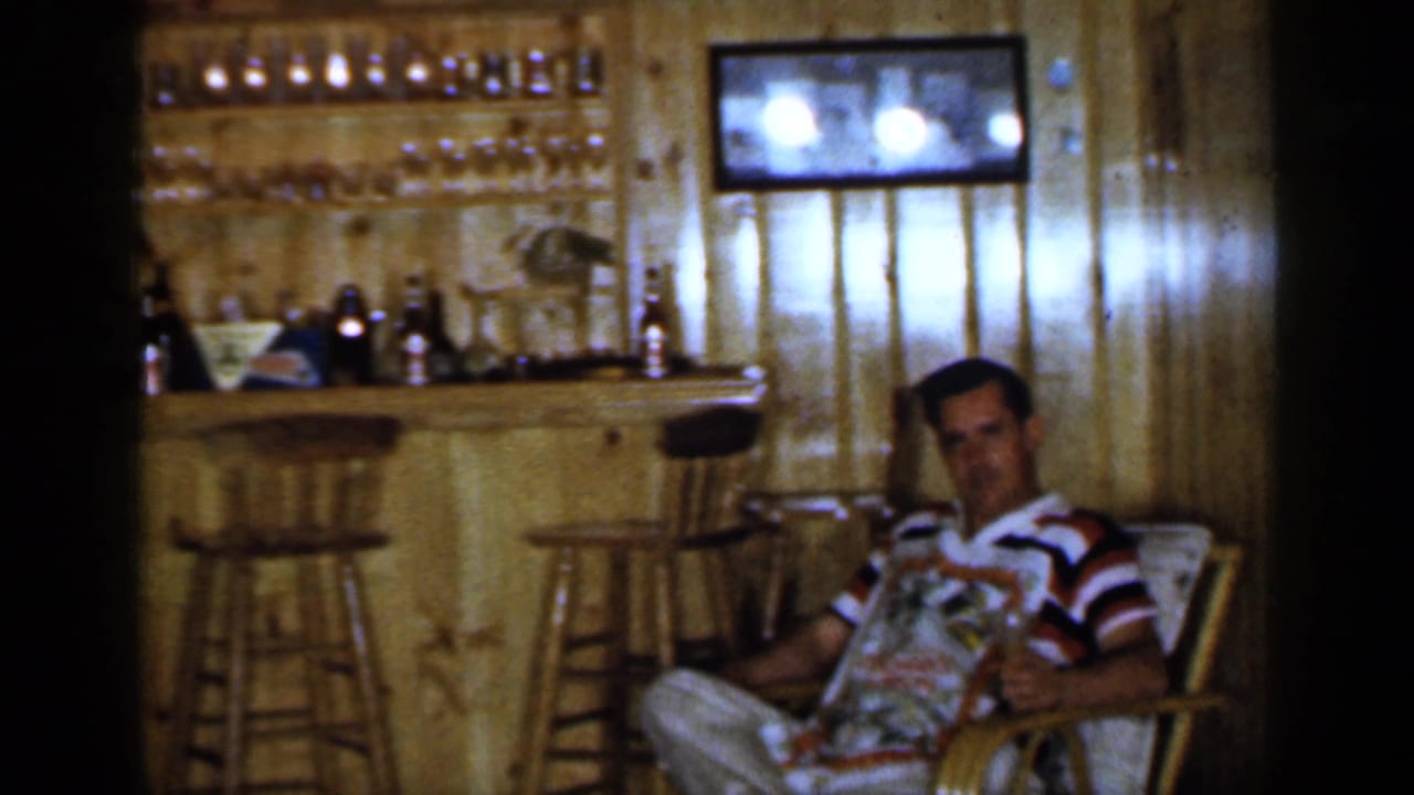 Man sits in a chair holding reading material beside a wood bar and wall shelves.