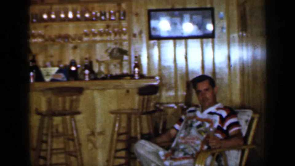 A man in a striped shirt appears to read or hold a magazine near a home bar in this 1965 Long Island interior.
