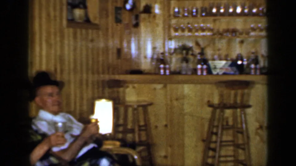 Home-movie frame from a private Long Island interior showing an older man seated near a lamp with a wood bar behind him.