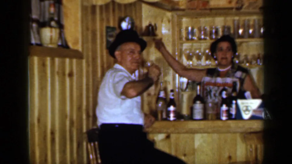 1965 Long Island home-movie frame showing an older man and woman toasting at a wood-paneled home bar with bottles visible.