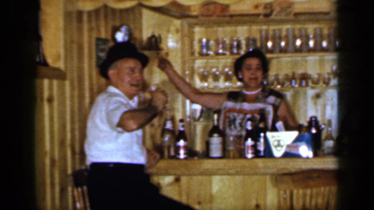 Man in a black hat and woman behind a wooden bar raise glasses together.