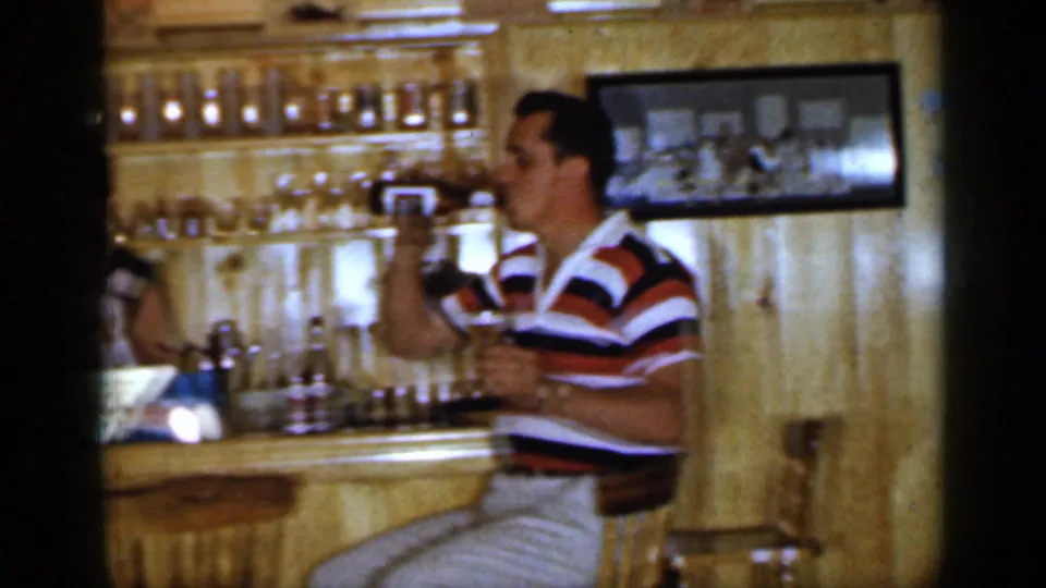1965 Long Island home-movie frame showing a man in a striped polo seated at a home bar and drinking from a glass.