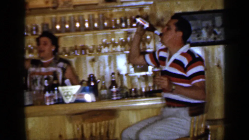 1965 Long Island home-movie frame showing a man drinking from a bottle near a woman behind a wooden home bar.