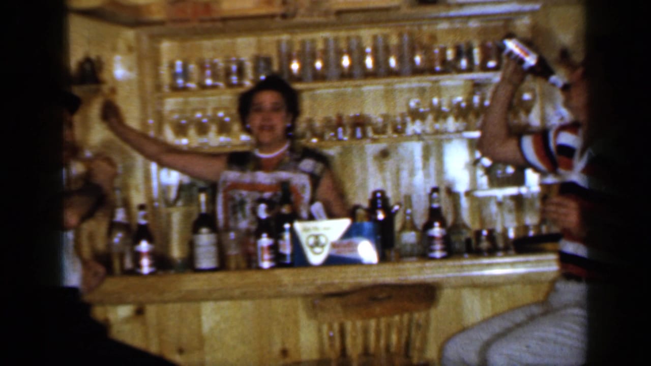 Woman behind a wooden bar raises her arms while beer bottles and glassware sit on the counter.