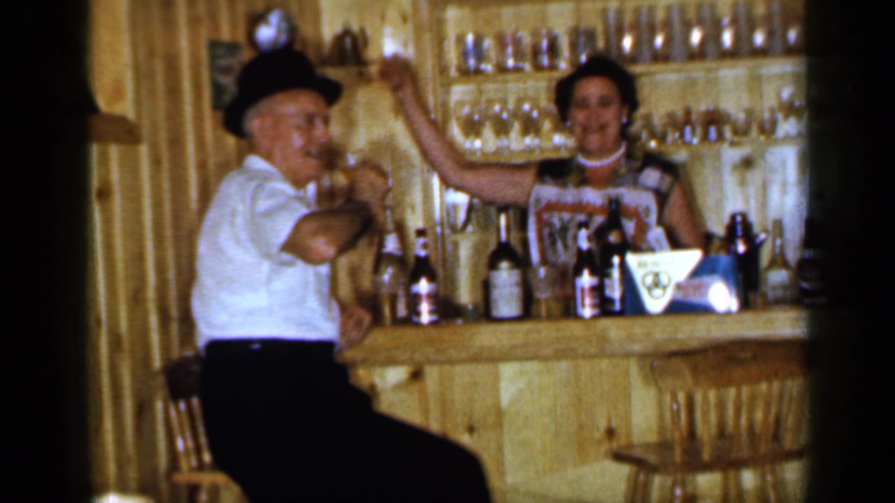 Man in a dark hat sits at the bar while a woman behind the counter raises one arm.