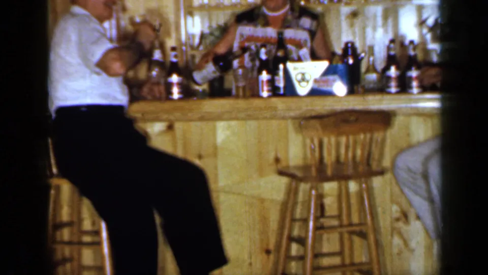 1965 Long Island home-movie frame of a man seated at a wooden bar, with beer bottles, stools, and a woman partly visible behind the counter.