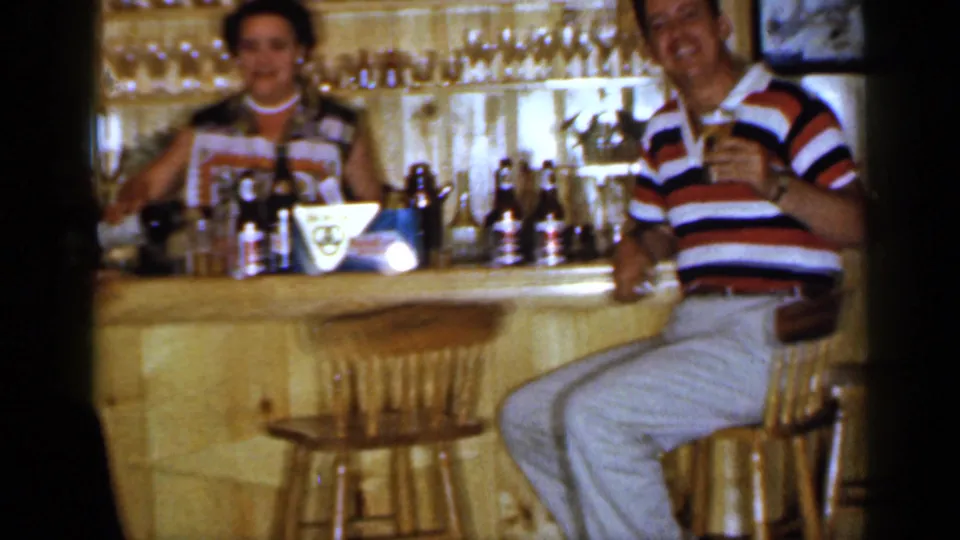 1965 Long Island home-movie frame of a smiling man in a striped polo at a wooden bar with a woman, bottles, and glassware behind him.