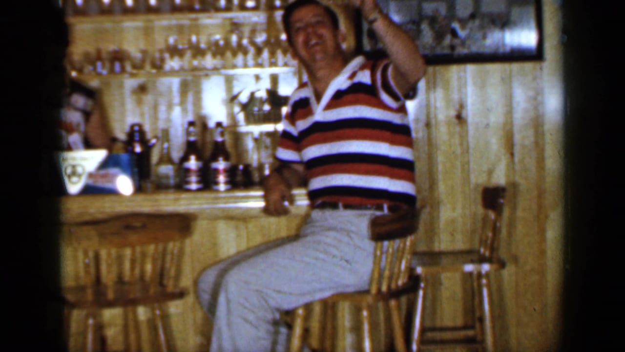 Man in striped polo raises a drink while seated at a wooden bar.