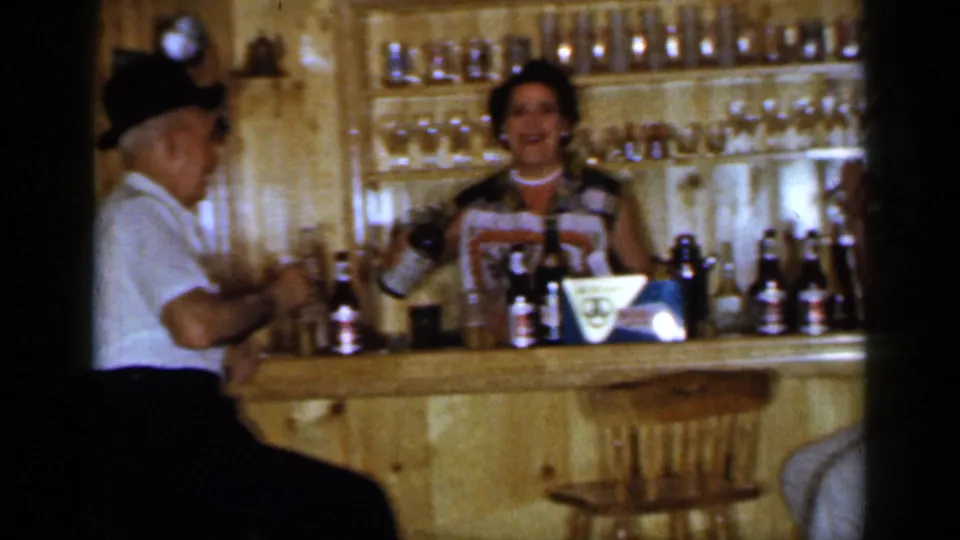 1965 Long Island frame with a man in a white shirt and black hat at the wooden bar as a woman stands behind bottles and glassware.