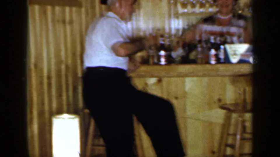 1965 Long Island home-movie frame showing a seated man at a rustic bar, with shelves of glassware and bottles in the background.